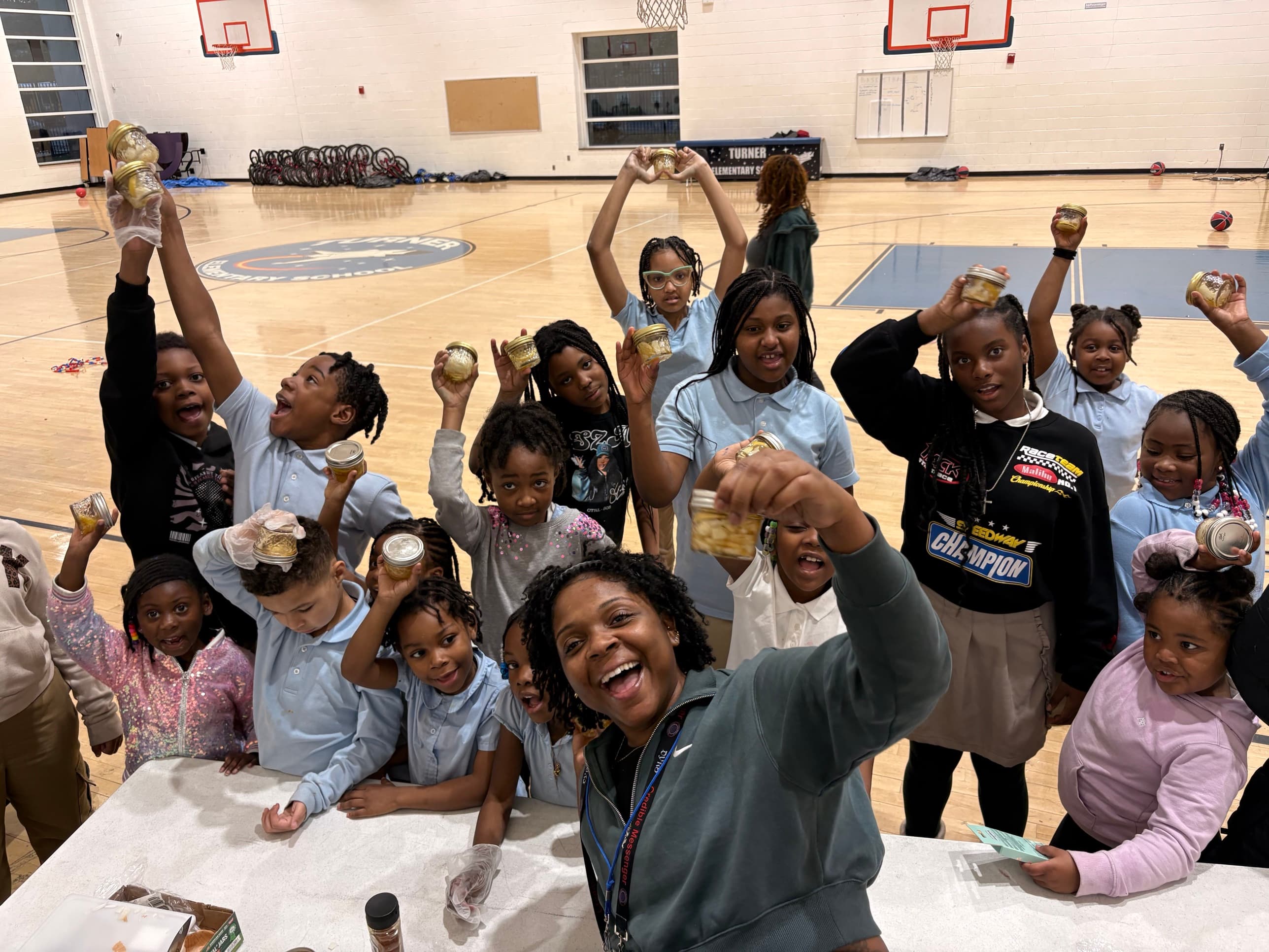 A smiling woman takes a group photo with about twenty children in a gym, many raising small jars they made.