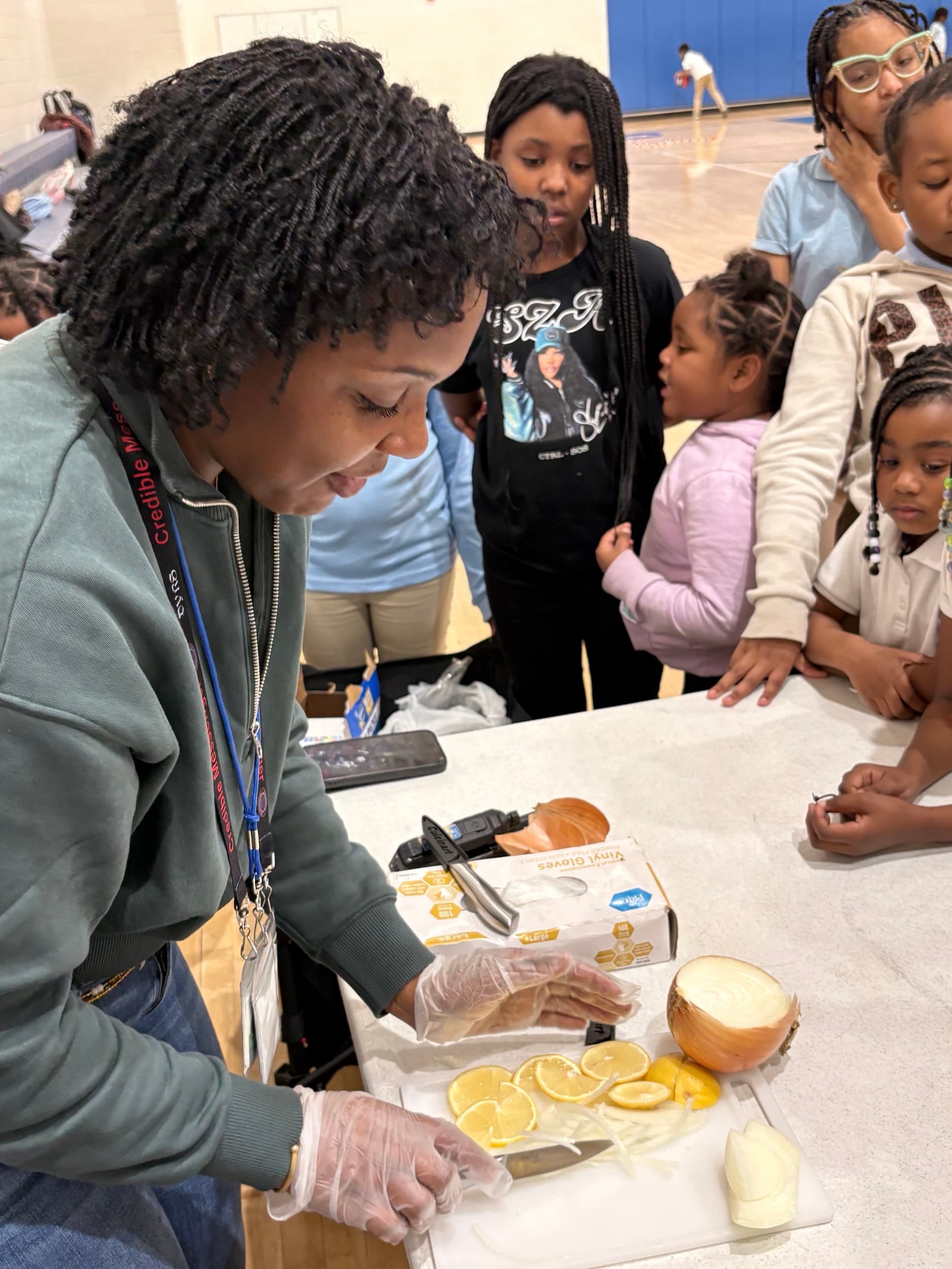 A woman slices lemons and onions at a table while a group of children watch closely.